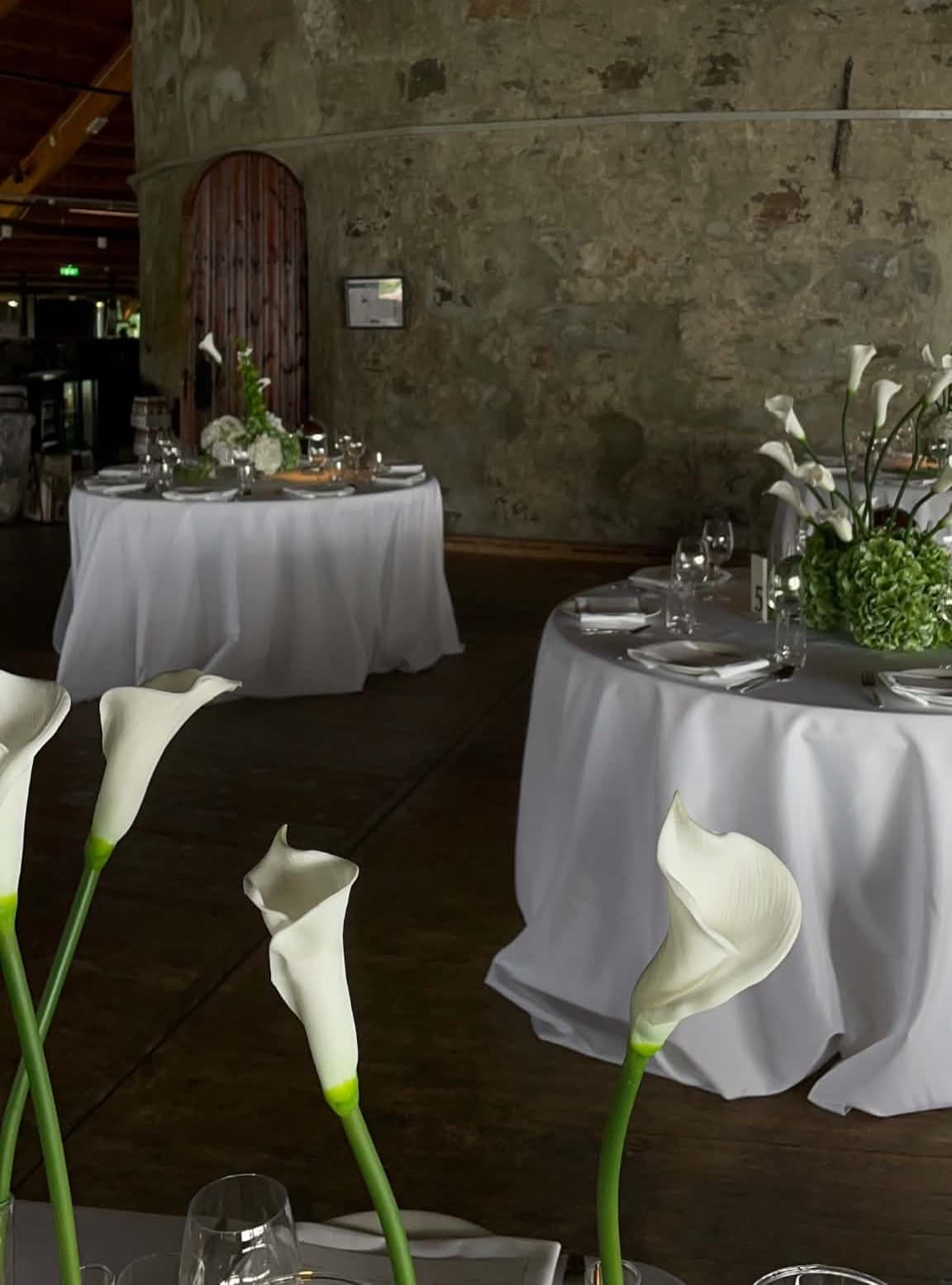 Runde festbord med hvite duker og blomster i et lokale med steinvegger
